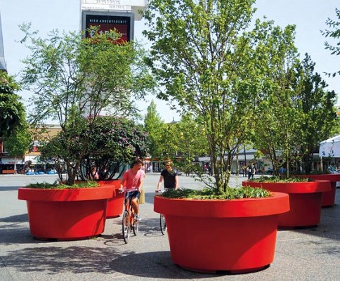 4 large red planters with trees green trees growing. Two cyclists travel between two of the planters