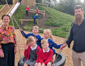 Adults and children pose on a backyard swing set: two adults stand beside three children seated together on a tire swing, smiling on a sunny day.