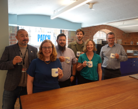 A group of people standing behind a bar, smiling and holding mugs, with the word 'Patch' in the background