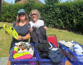 Two smiling women pose outdoors beside carts filled with clothing and bags; one holds green shoes, with hedges and a sunny lawn behind them.