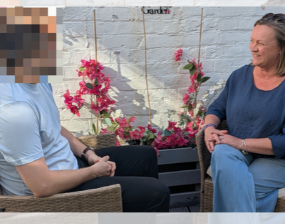Two people sit facing each other beside pink flowering vines on a trellis against a brick wall, sharing a calm conversation outdoors.