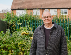 A smiling man with glasses and gray hair stands in a lush community garden, with a green fence and brick houses behind him, conveying warmth and ease.
