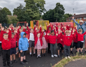 A cheerful school group in red uniforms gathers on a playground, raising their hands beneath a cloudy sky, with colorful climbing frames and trees behind them.
