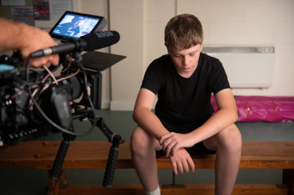 A boy in black sports kit sits downcast, as a camera films him