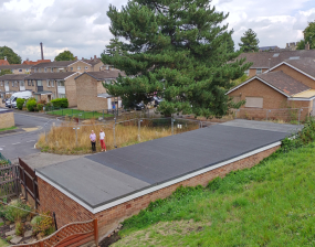A view across some garage roofs to a residential area of homes and boarded buildings with a large tree in the middle.
