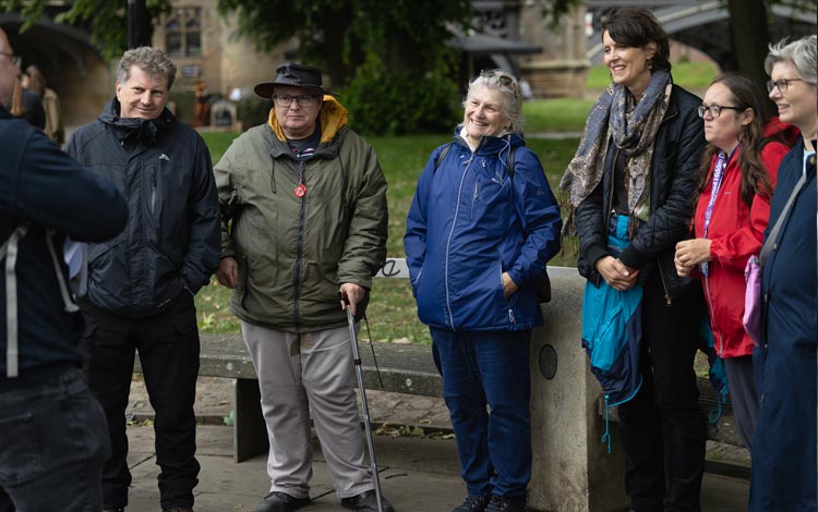 A group of people standing together on a riverside path in York, with one person using a walking stick and a stone bridge visible in the background.