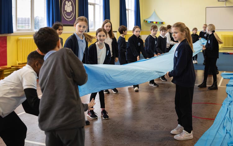 Children in a school hall working as a group to hold up a strip of blue fabric, pretending it's a river.