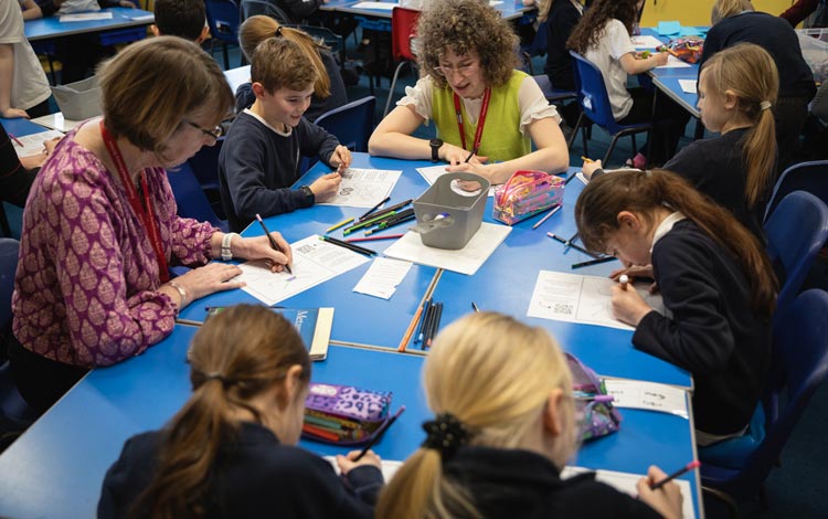 Children and an adult working together at tables in a classroom, drawing and completing activity sheets with pens and pencils.