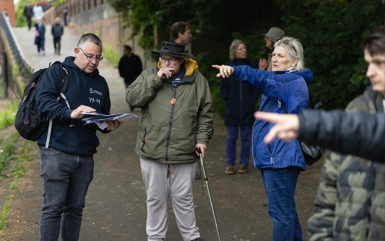 A small group of people standing on a riverside path, talking together, while one person points ahead and another holds printed materials.