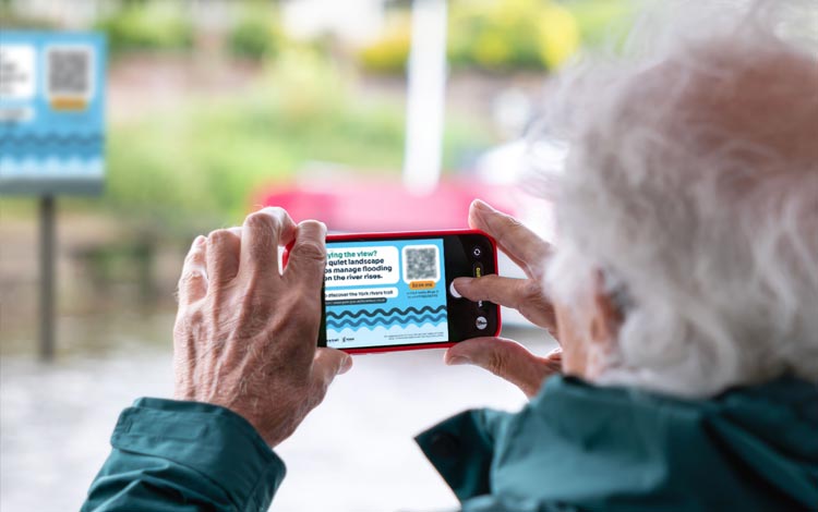 A person at the riverside, using a mobile telephone to scan the QR code on a signpost.