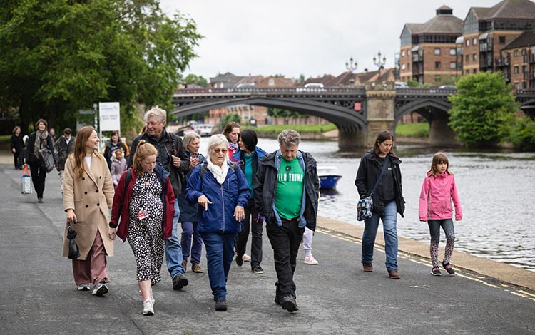 A group of people walking along a riverside path in York, with the river, bridge and historic buildings visible in the background.
