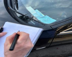 A hand holding a pen is poised over a notepad next to a Blue Badge displayed in a car window