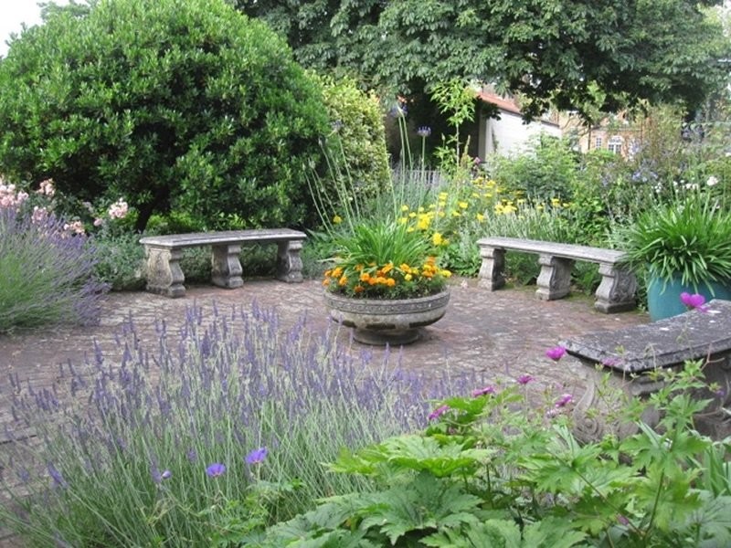 This is an example of what the community garden will look like, it shows a circular path of gravel surrounded by benches which have been evenly placed around a plant pot in the centre. The image shows lots of greenery around the space.