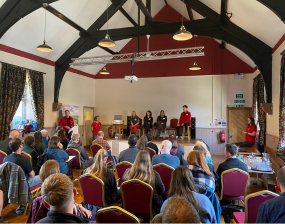 A room of people listening to speakers on a panel at the front of the room