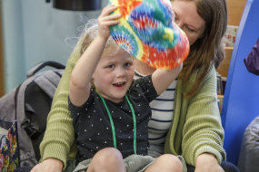 Photo of a young child sat on an adult's knee, waving a colourful hat