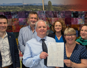 A group of six people stand smiling in front of a colorful quilt mural. One person in the center holds up a document, suggesting a celebration or achievement.