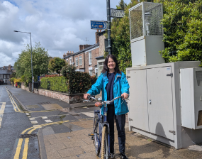 A women dressed in blue, holding a bike, standing next to an air quality monitor station in York