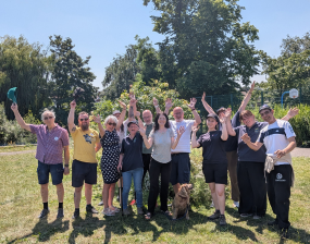 Group of people standing outdoors on a sunny day, smiling and waving with a dog sitting in front of them on green grass.