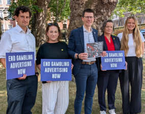 Five people standing outdoors holding signs that say 'END GAMBLING ADVERTISING NOW' in a campaign setting.
