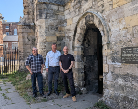 Three men standing beside an ancient stone archway entrance on a sunny day in a historic urban area. / Plaque detailing St. Leonard's Hospital history, built by John Romanus in 1255 with a vaulted crypt and chapel above.