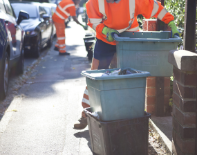 a waste worker in a bright orange uniform collects a recycling bin on a sunny street, conveying industriousness and community service.