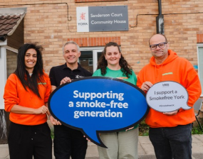 Four smiling community members pose outside Sanderson Court Community House, holding signs supporting a smoke-free generation and Smokefree York.