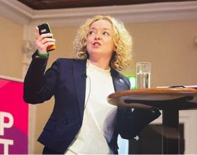 a blonde haired woman in a smart outfit is presenting at a lectern