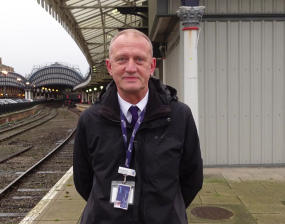 A person in a black jacket stands at a train station platform with tracks and an arched roof visible. The setting is overcast, and the atmosphere is calm.