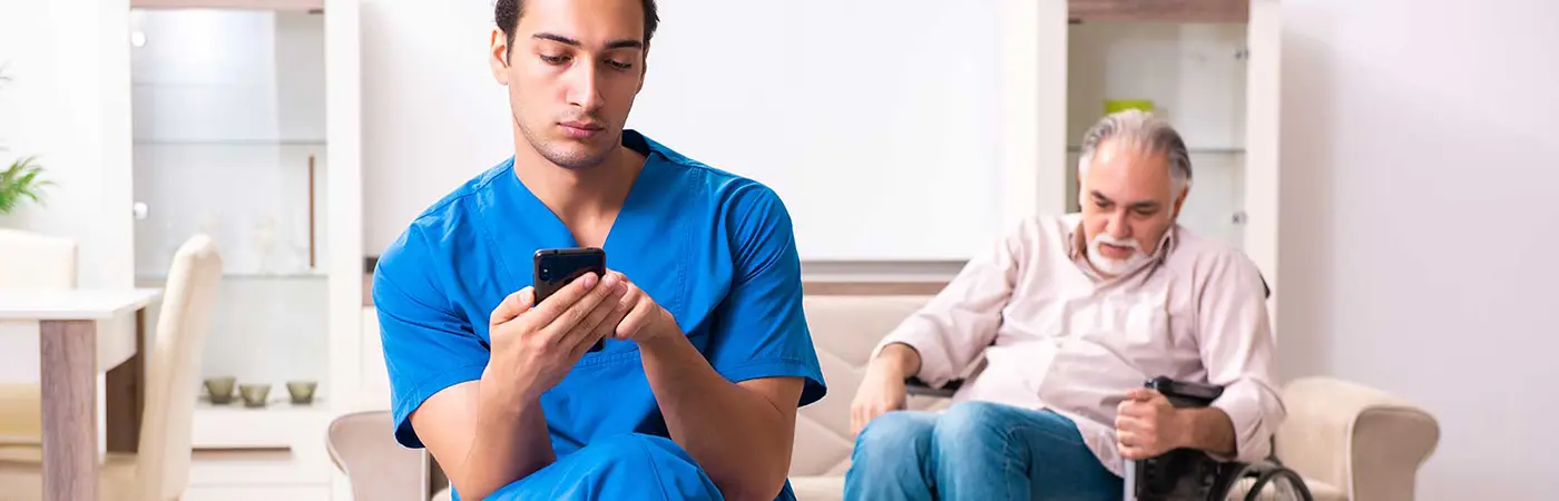 A male nurse sits in a chair using his mobile phone whilst an elderly patient calls out for help.