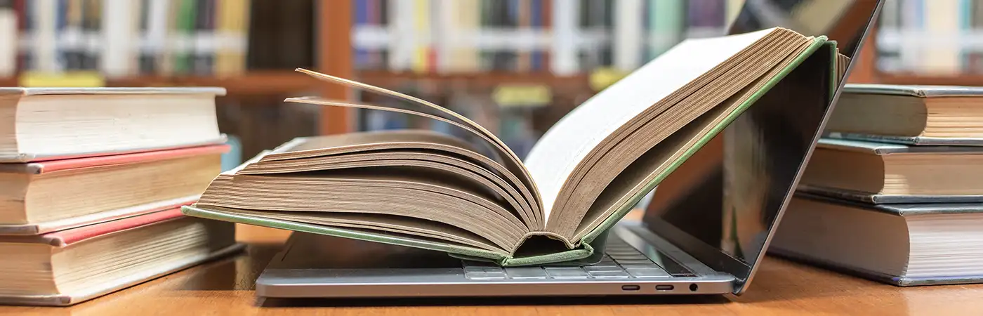 A pile of text books surrounds a laptop PC. There is an open book lying on the laptop. There are rows of books in the background.