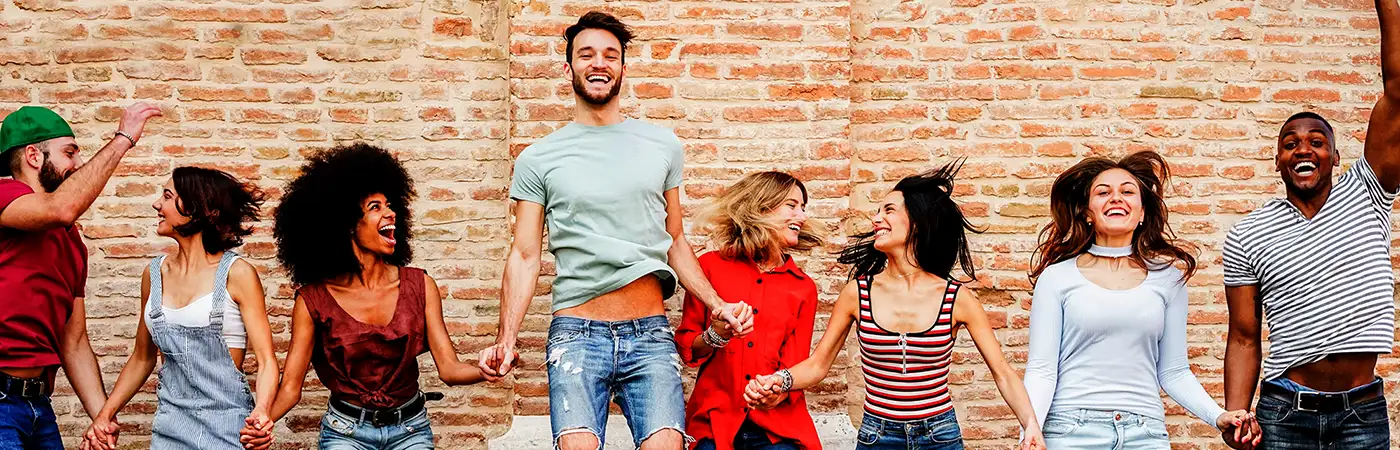 A group of young people are all jumping in the air and laughing. There is a brick wall behind them.